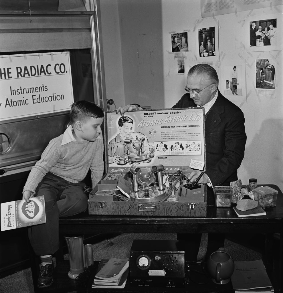 An unspecified young boy alongside an unspecified elderly man, in front of them is a 'Gilbert U-238 Atomic Energy Laboratory,' with a poster reading 'The Radiac Company, instruments for the uranium prospector,' at the Atomic Center (formerly known as the Radiac Company), one of the first companies to offer a variety of Geiger and scintillation counters to the general public, in New York City, New York, 19th February 1952. The toy lab was produced by the A C Gilbert Company in 1950 for children to create and watch nuclear and chemical reactions using radioactive material. (Photo by Canswell/Graphic House/Archive Photos/Hulton Archive/Getty Images)