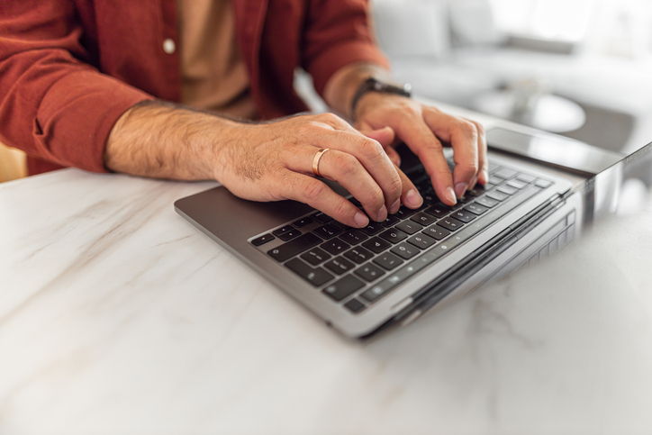 A close-up shot of adult man's hands using a laptop and typing on a the keyboard while working remotely on the job.