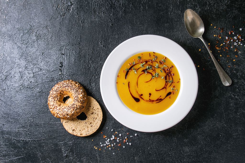 Plate of vegetarian pumpkin carrot soup decorated by balsamic vinegar and thyme served with herbs. spoon. bagel bread. chili pepper over black texture background. Flat lay. space. (Photo by: Natasha Breen/REDA/Universal Images Group via Getty Images)