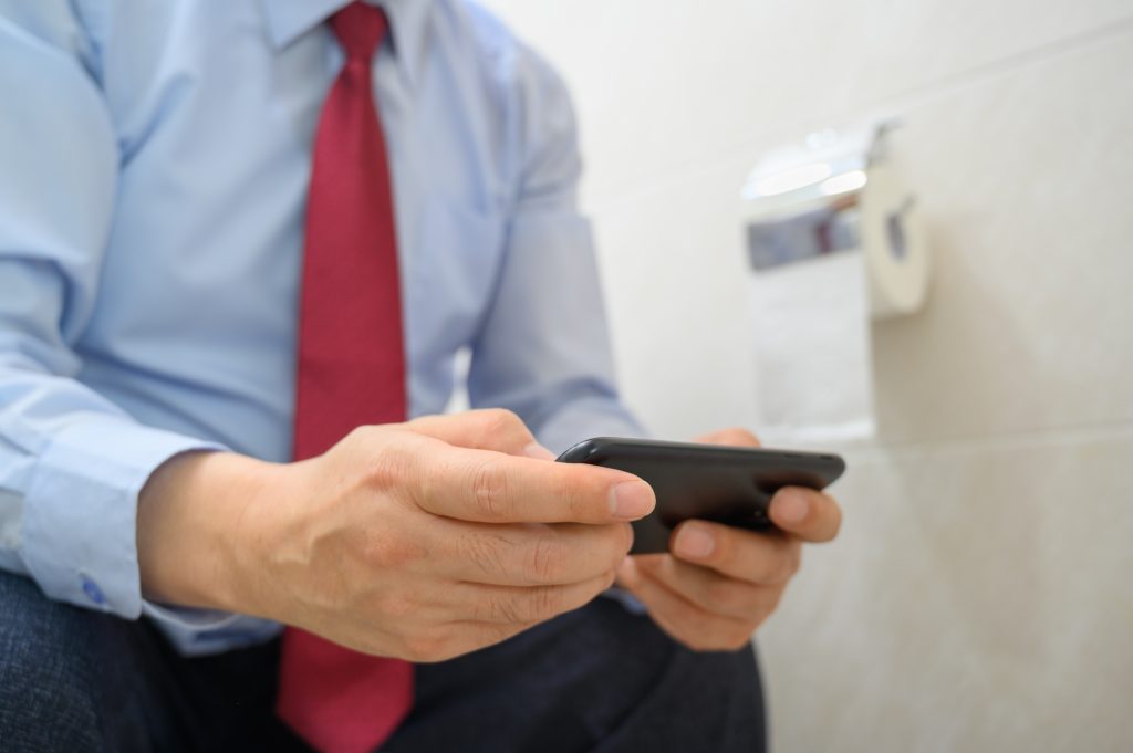 Businessman sitting on the toilet in the bathroom and using a smartphone.